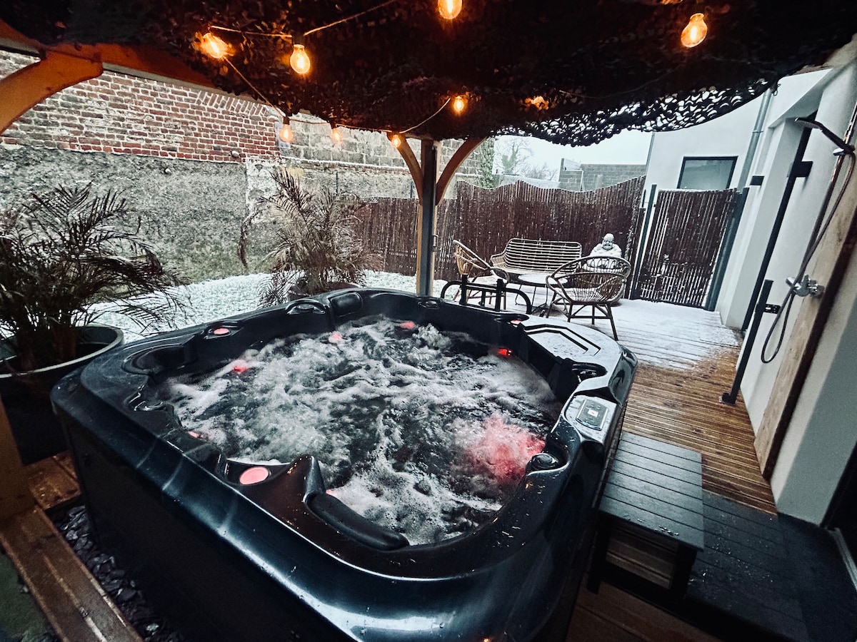 A covered private spa is showcased in an outdoor setting, featuring bubbling water and soft lighting from overhead pendant lights. Wooden decking surrounds the spa, accompanied by comfortable seating and privacy screens that create a serene atmosphere.