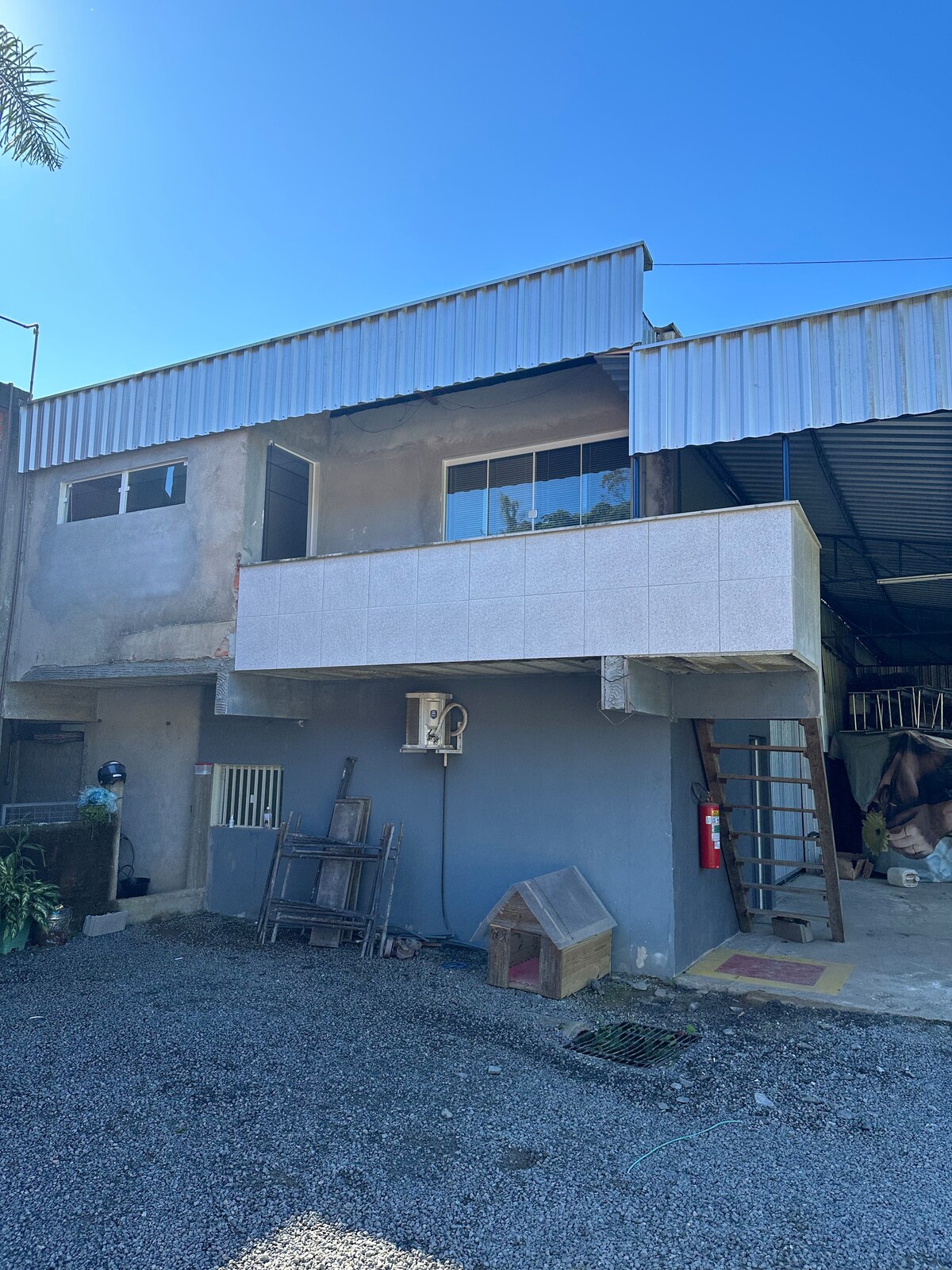 The two-story building is characterized by a mix of materials, featuring a prominent balcony with a large window above a shaded area. A stairway leads to the upper level, while outdoor spaces are filled with gravel and greenery.