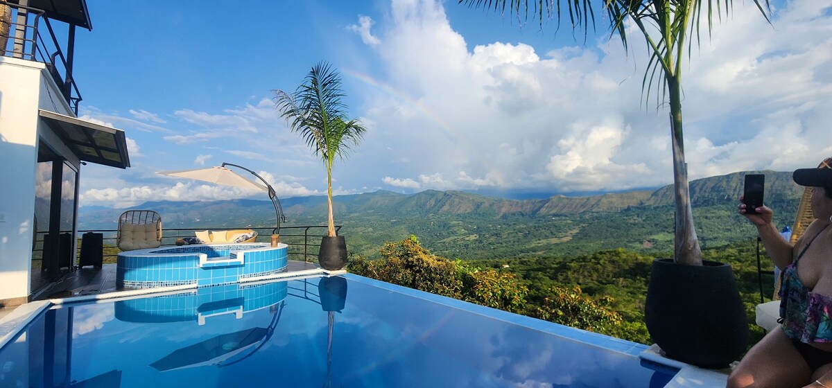 An outdoor pool area is set against a backdrop of rolling green hills and a colorful rainbow in the sky. Tall palm trees are positioned near the pool, which features a blue tiled edge and is surrounded by lush vegetation. Comfortable lounge chairs are arranged for relaxation.