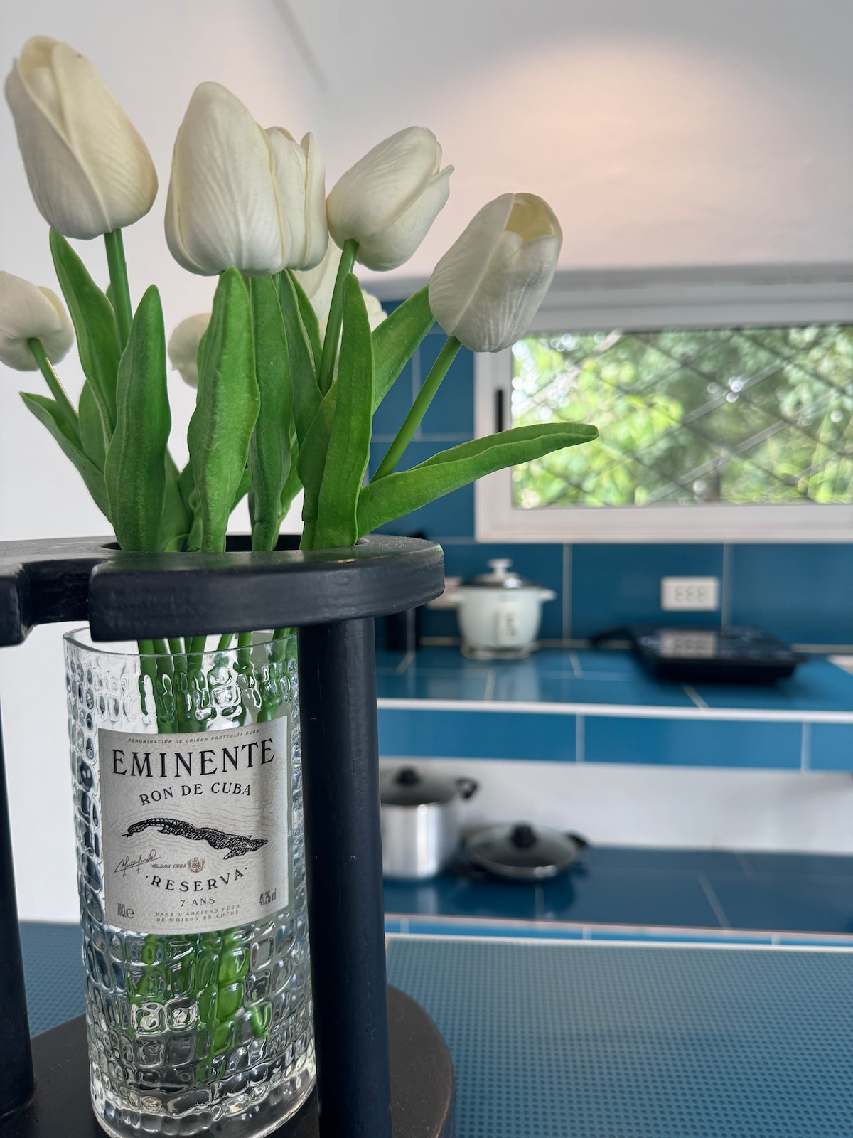 A clear glass vase holds white artificial tulips, positioned on a dark surface. Behind it, a bright kitchen area features blue-tiled walls and modern appliances, including a kettle and cooking pots, with natural light entering through an open window.