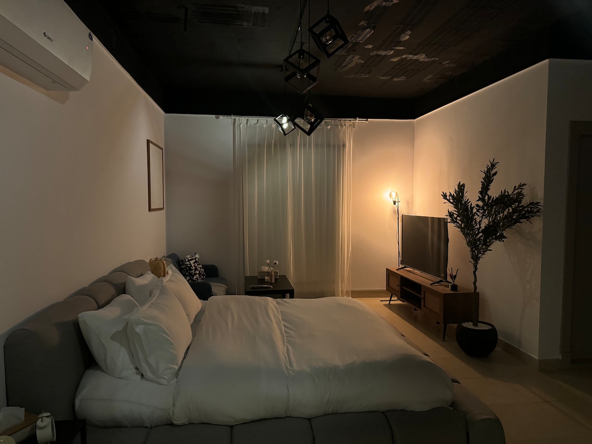 A spacious bed is positioned in the corner of the room, draped in soft white linens. A seating area with two glasses on a table is visible nearby. A smart TV sits on a wooden console, and a plant enhances the natural feel of the space.