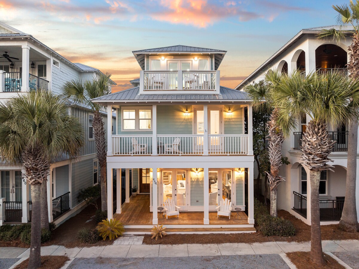 The exterior of a two-story beach house is showcased, featuring a light blue facade and multiple balconies. White rocking chairs are positioned on the front porch. Palm trees line the walkway, and the surrounding architecture reflects the coastal neighborhood's style.