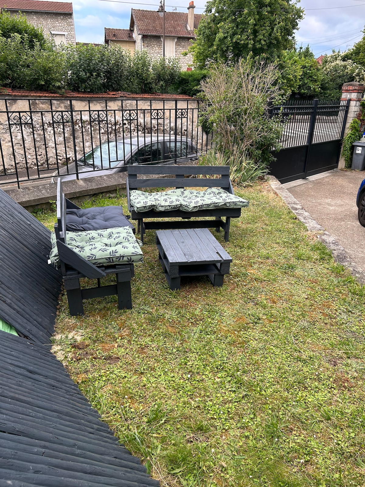 An outdoor garden area features a seating arrangement consisting of a dark wooden bench and two matching chairs, adorned with green patterned cushions. A small table complements the seating, creating a cozy space for relaxation. Lush greenery surrounds the area, contributing to a serene environment.