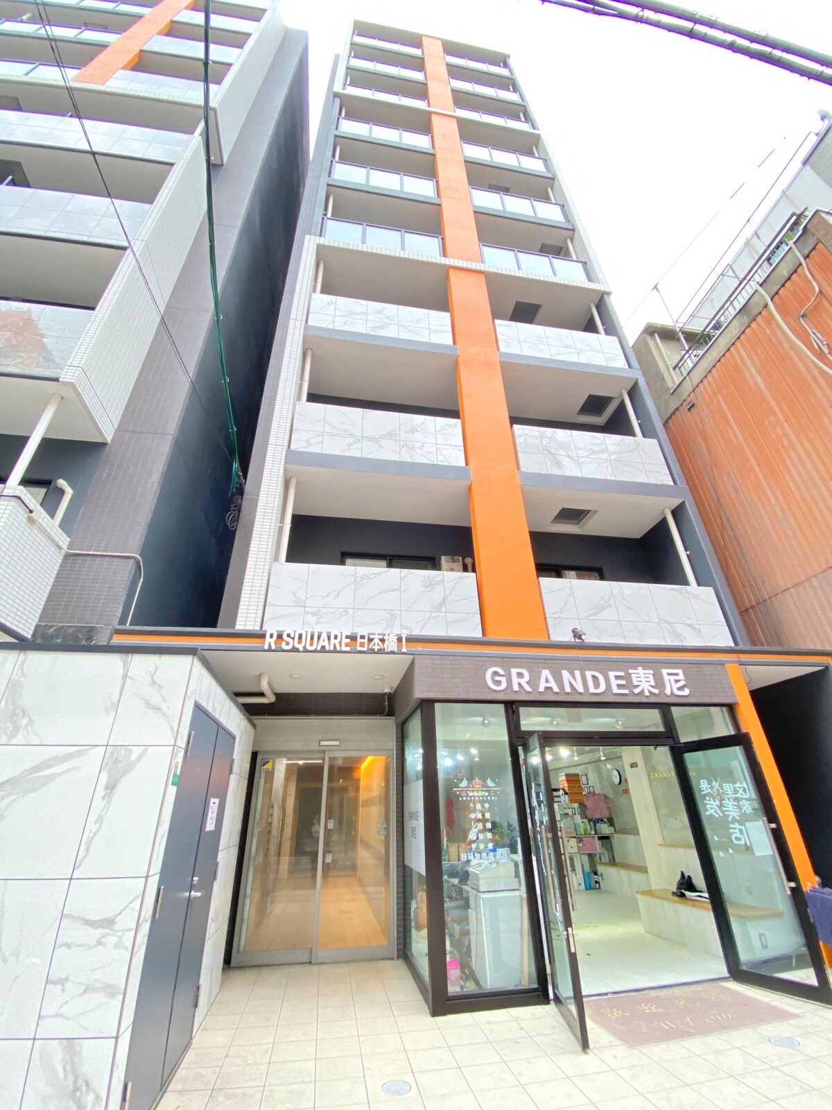 A modern apartment building with an eye-catching orange and gray facade is highlighted. The entrance features large glass doors, welcoming guests into a well-lit lobby area. Balconies are visible on the upper floors, contributing to the building's contemporary architecture.