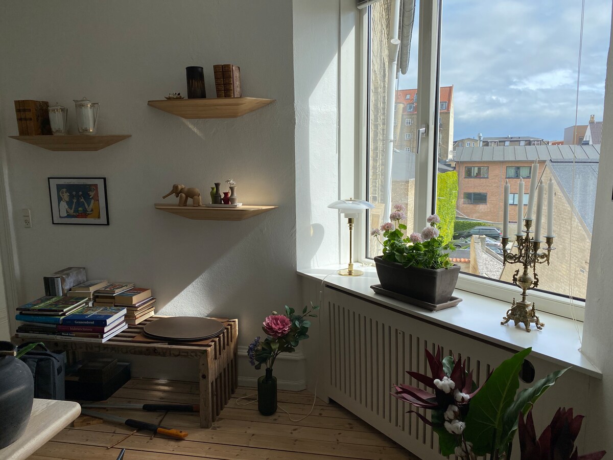A bright corner of the room features a large window with a view of the outside. Shelves are mounted on the wall, displaying decorative items and books. A potted plant sits on the windowsill, accompanied by a lamp and a decorative candelabra.