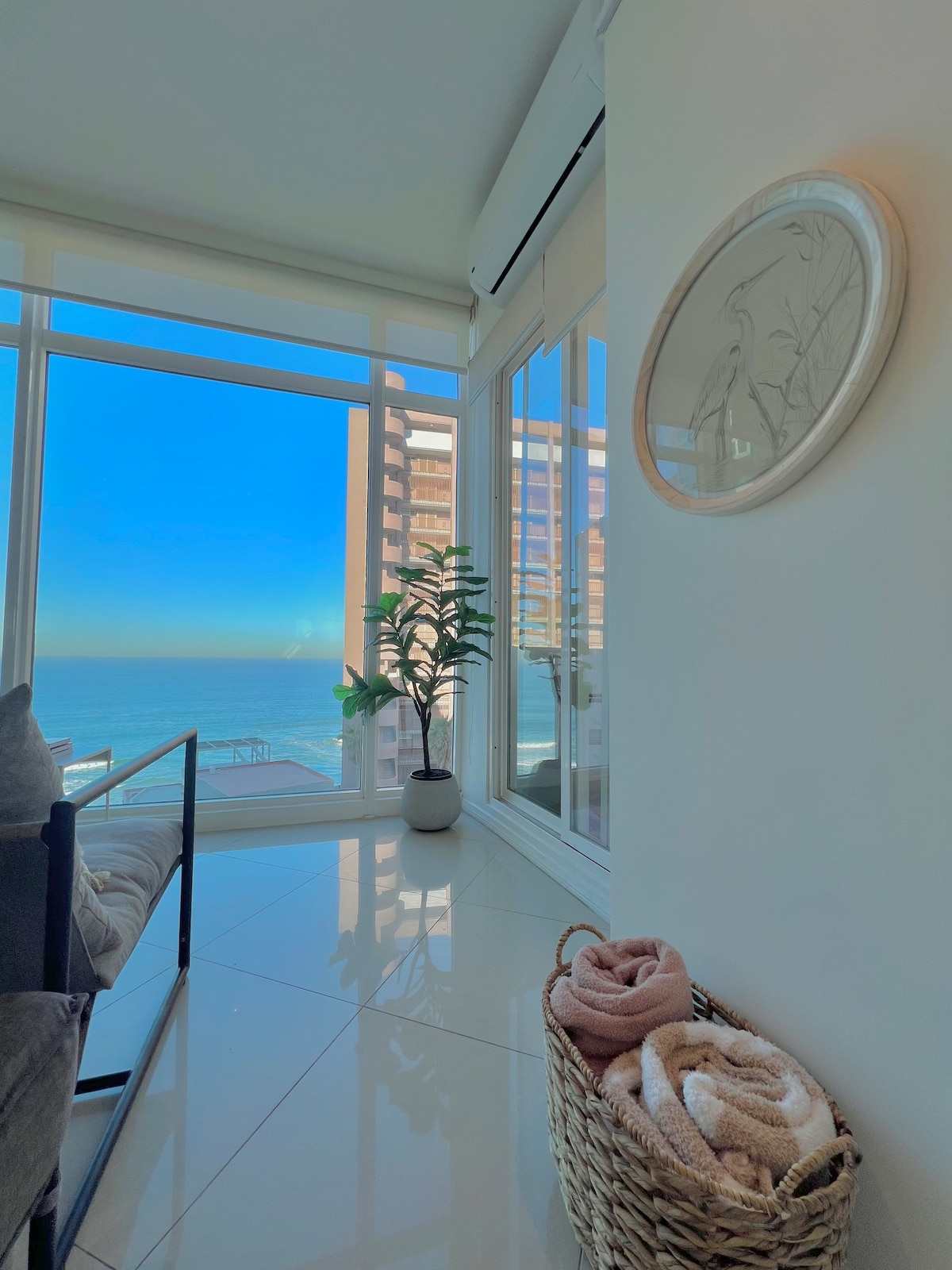 A corner of a modern living space features a cozy seating area with a view of the ocean through large glass windows. A potted plant adds a touch of greenery, while a woven basket holds neatly arranged towels. Soft natural light illuminates the room.