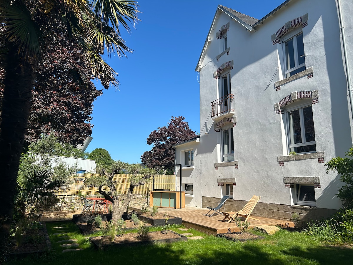 The house features a white exterior with multiple windows and a balcony on the upper floor. A spacious garden area with grass is complemented by wooden decking and sun loungers. Surrounding greenery includes palm and olive trees, enhancing the outdoor space.