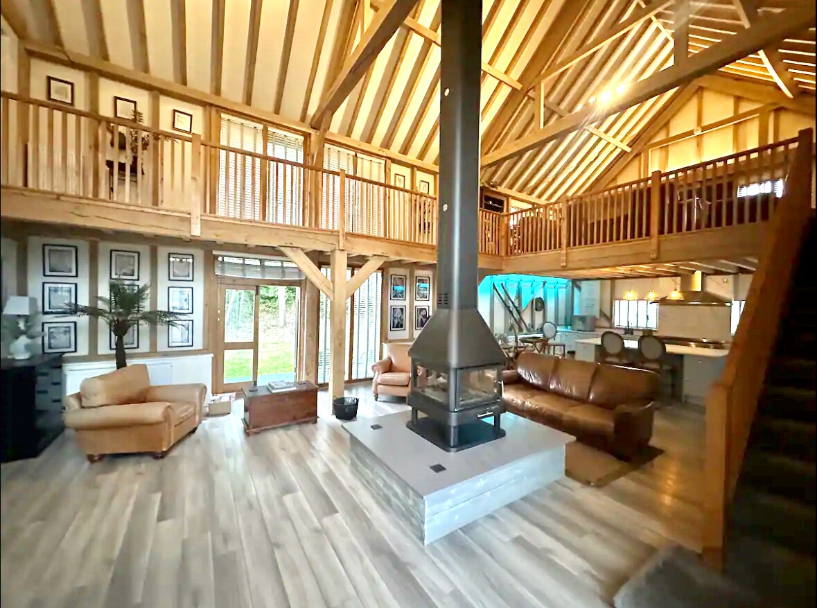An open-plan living space showcases high wooden beams and a modern fireplace at the center. Comfortable leather sofas are arranged around the fireplace, while large windows bring in natural light. Wooden accents and framed photographs enhance the rustic charm of the room.
