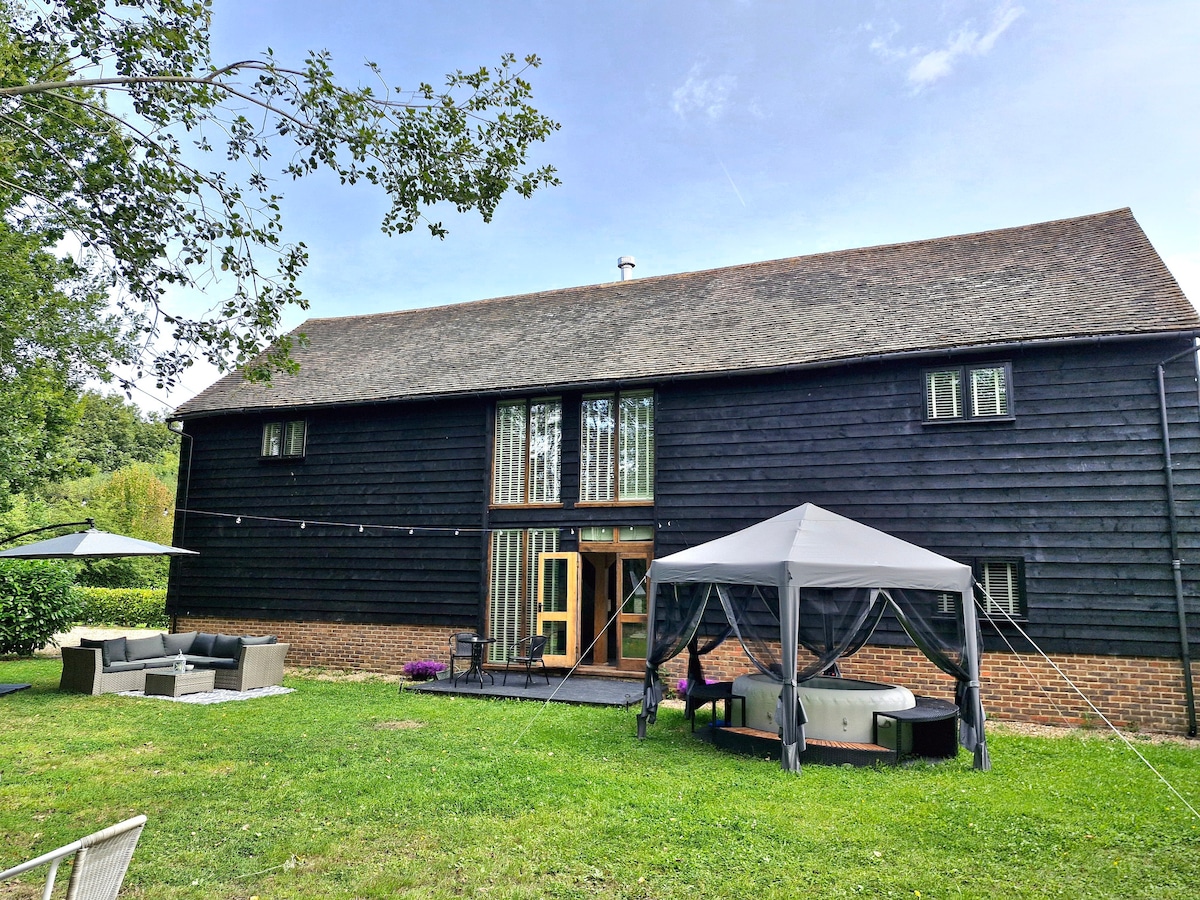 The exterior of the barn showcases a dark wooden façade complemented by large windows. The lawn features a gazebo, a hot tub, and a comfortable outdoor seating area. A patio extends from the barn, seamlessly connecting indoor and outdoor spaces.