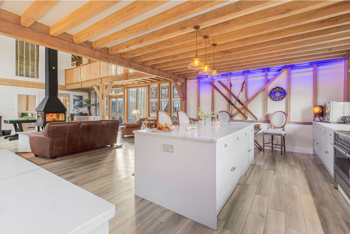 An open-concept kitchen and living area is showcased with exposed wooden beams and modern finishes. A spacious island with white countertops provides a central gathering spot. The room is illuminated by pendant lights, while a cozy fireplace adds warmth to the inviting atmosphere.