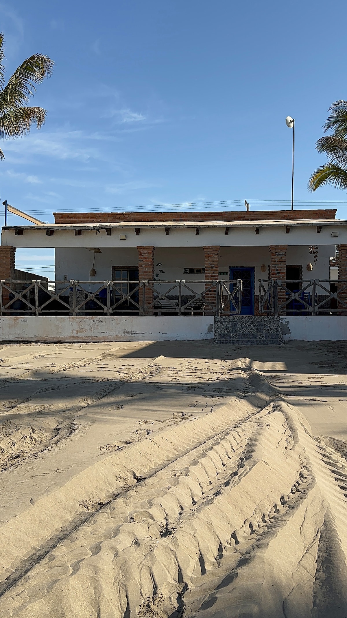 A beachfront property is presented with a simple design. The exterior features a covered porch with wooden railings and proximity to the sandy area. Palm trees are visible nearby, and a blue door serves as the main entrance, suggesting a casual and welcoming environment.