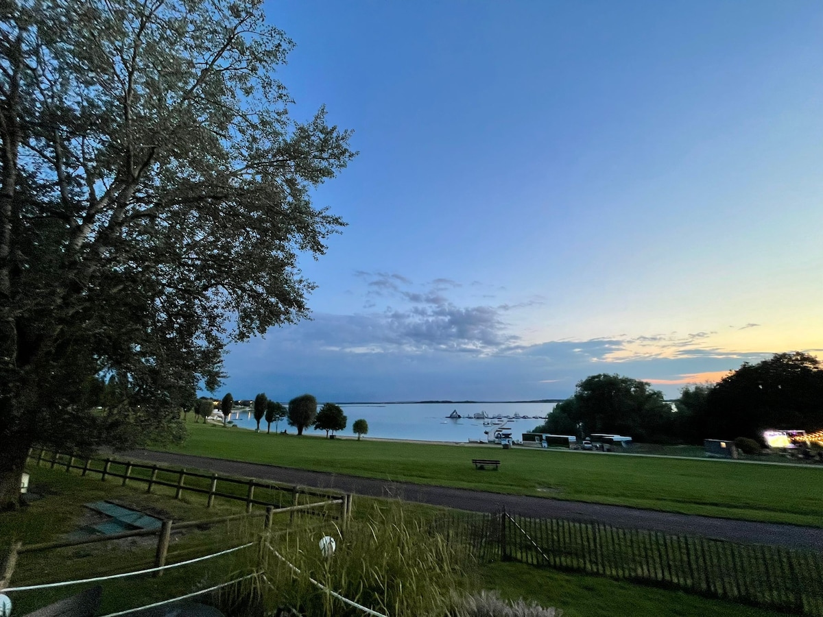 A serene lakeside view is captured at twilight, showcasing calm waters reflecting the soft evening sky. Silhouetted trees frame the scene, while boats can be seen gently bobbing on the lake. A grassy area leads to the water's edge, inviting relaxation.