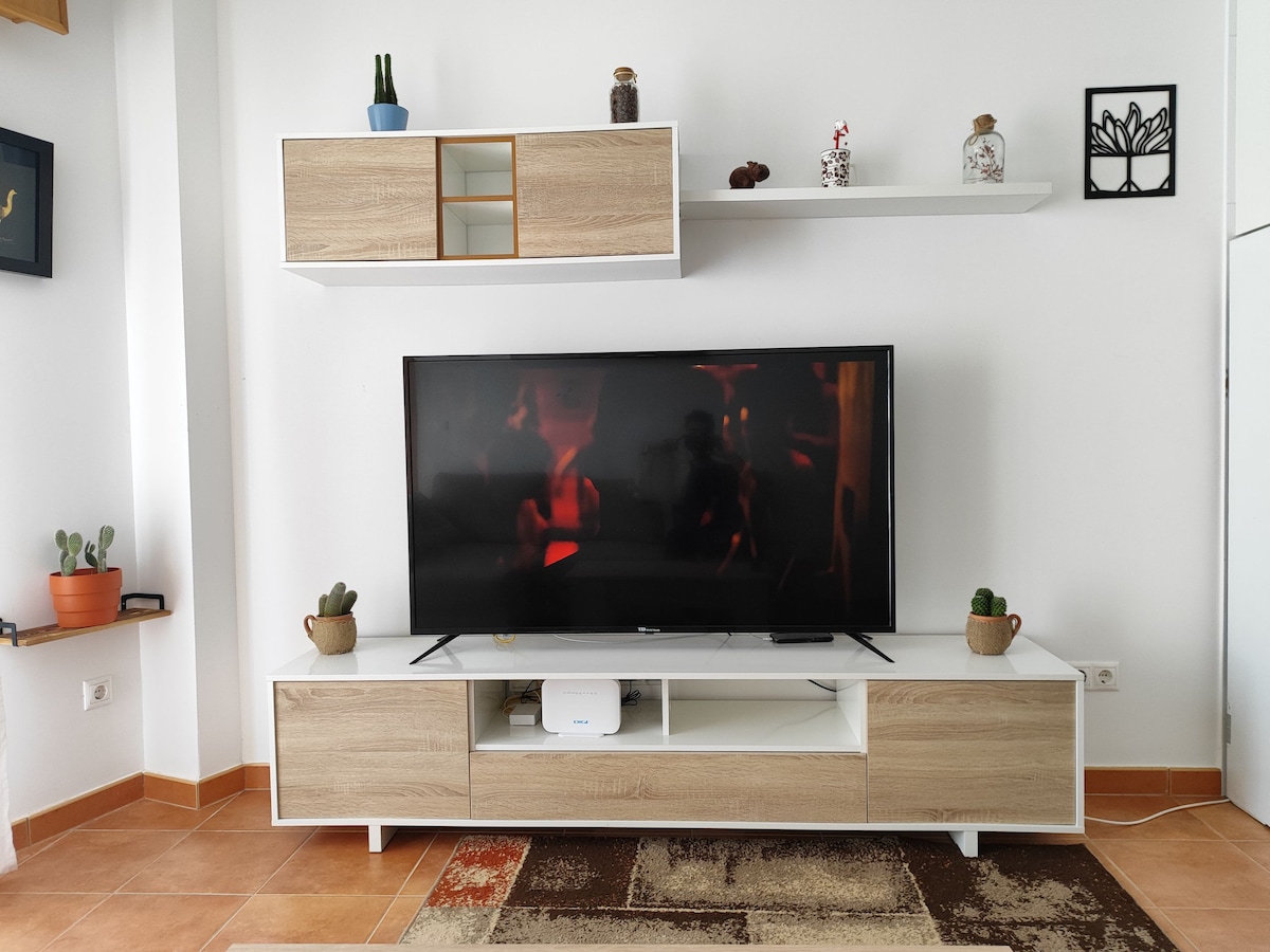 A modern entertainment area features a large flat-screen TV positioned on a light wooden console. Above, a mounted shelf displays decorative items and small potted plants. The neutral wall color and warm tile flooring create an open and relaxed environment.