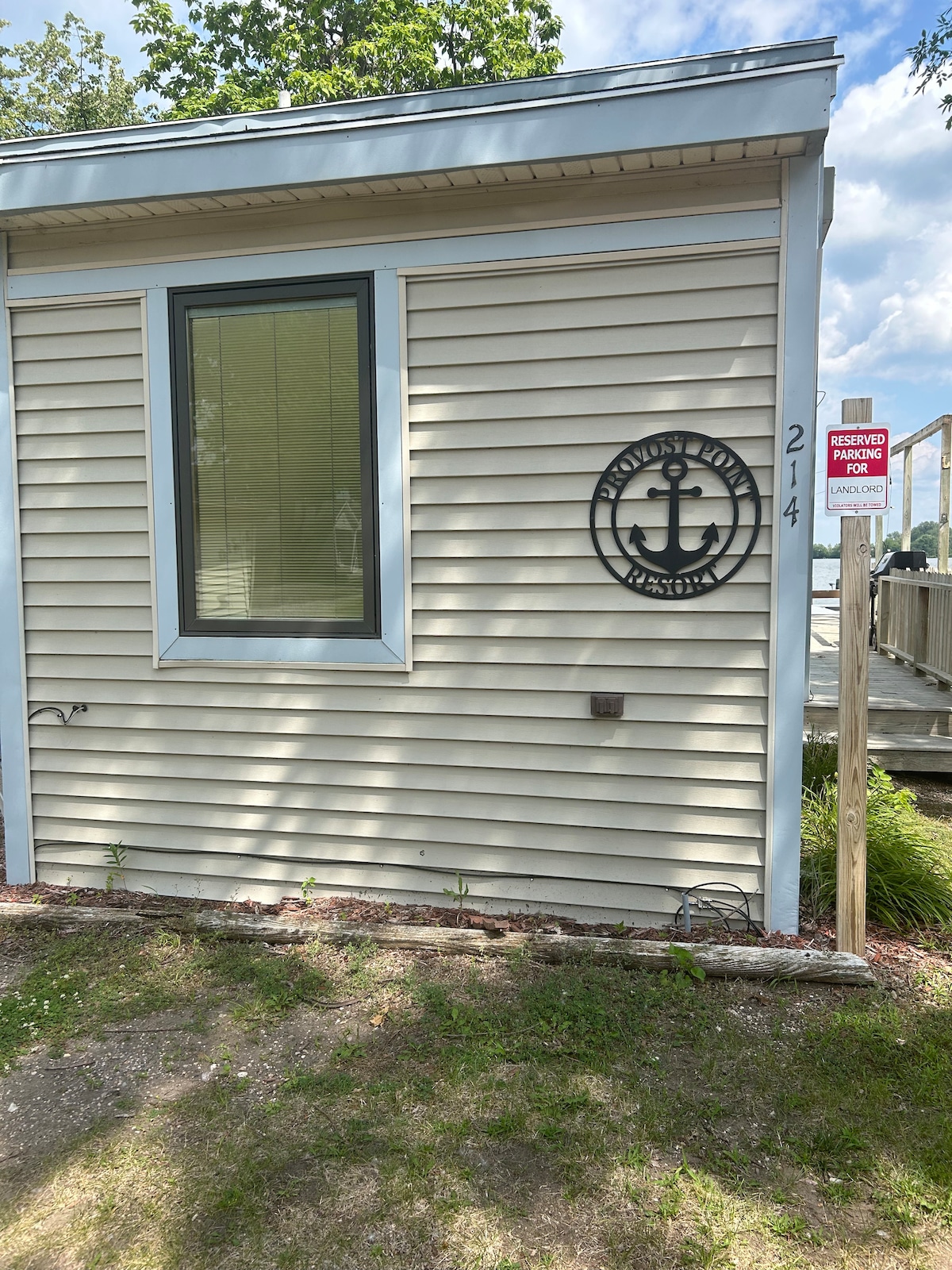 The exterior of the property features light beige siding and a large window, providing a view of the surroundings. An anchor-themed sign is mounted on the wall, indicating the property number. The lawn area is neatly maintained, with some greenery present.
