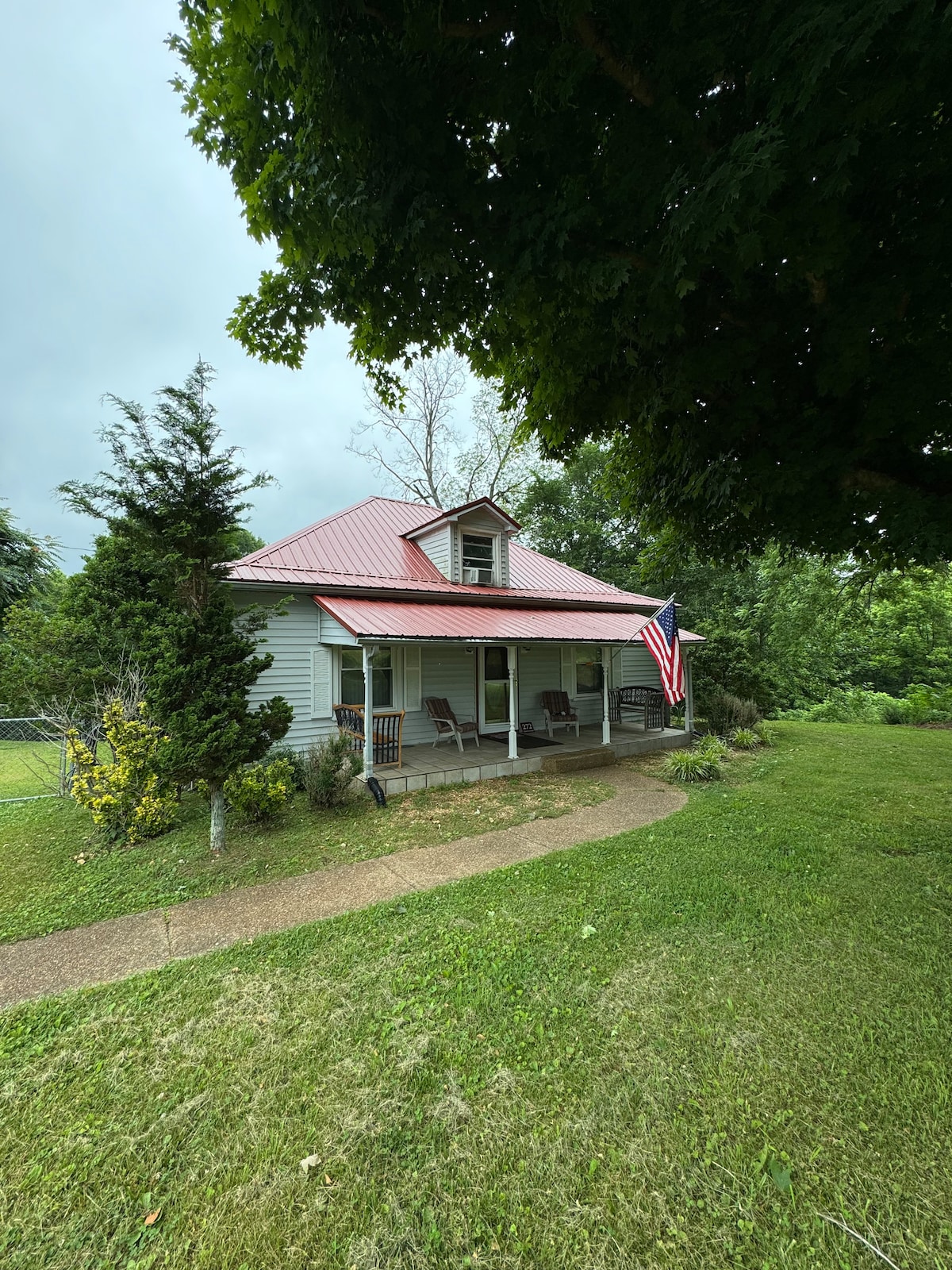 A charming home with a red metal roof is surrounded by greenery. A front porch features rocking chairs, inviting relaxation. A pathway leads to the entrance, decorated with vibrant foliage and a flag gently swaying in the breeze, adding to the welcoming outdoor ambiance.