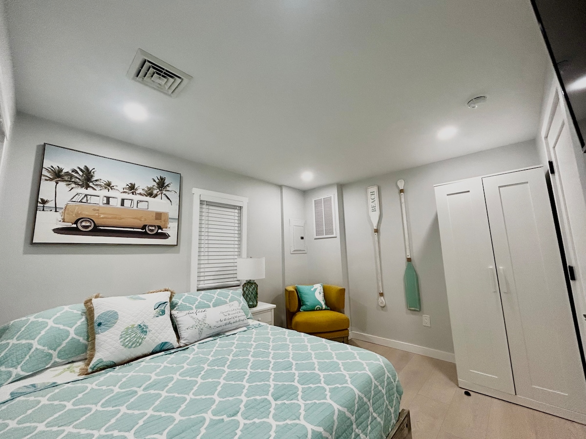 A bright and inviting bedroom is enhanced by coastal decor. A spacious bed with a patterned coverlet is paired with a small bedside lamp. A comfortable chair sits beside a closet, and a framed image of a vintage van adds personality to the space.
