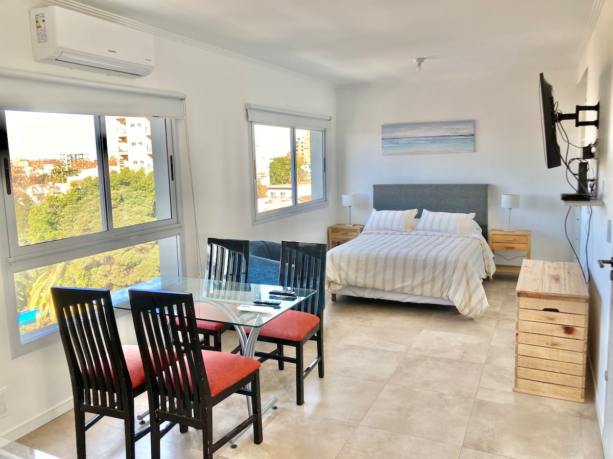 A bright, open living space is depicted, showcasing a spacious bed with neutral bedding. Four dining chairs surround a glass table. Natural light floods in through large windows, revealing views of the surroundings. A wall-mounted TV is visible, alongside a wooden console below.