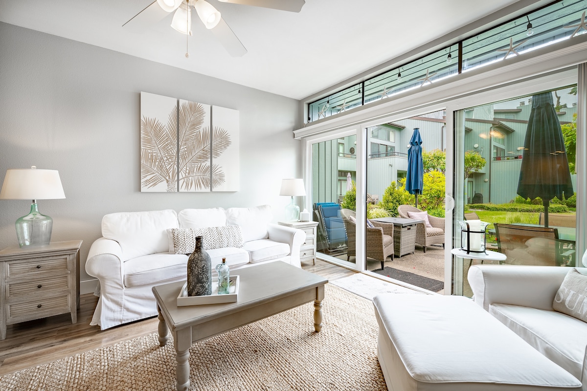A bright living area features a white sofa and a matching armchair, complemented by a neutral area rug. Large glass doors open to a patio area with green landscaping and outdoor seating. Two lamps provide soft lighting, enhancing the inviting ambiance.