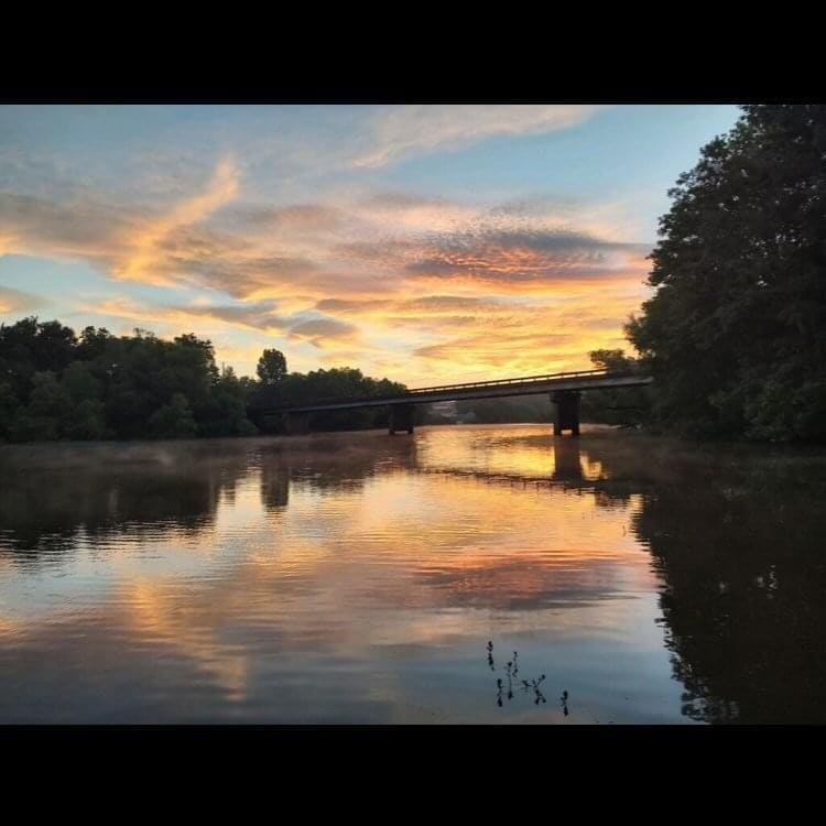 A peaceful scene of a river at sunset is visible, with clouds reflecting vibrant colors on the water's surface. A bridge spans the river, surrounded by lush greenery, while the calm water captures the hues of the sky, creating a tranquil atmosphere.