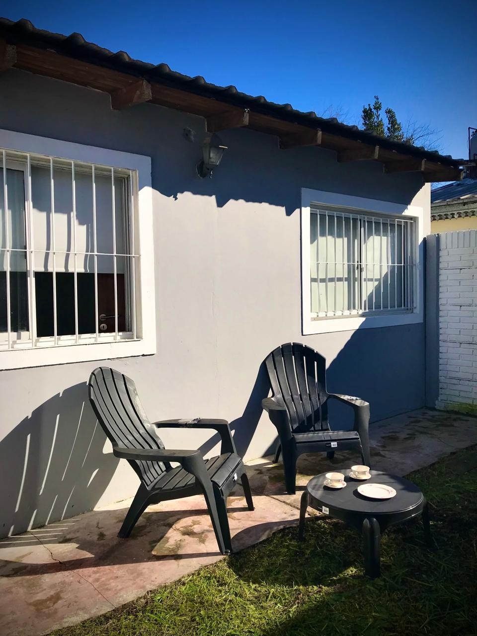 An outdoor seating area features two black chairs positioned on a paved surface. A small table holds three plates, and sunlight illuminates the space, highlighting the neutral-colored walls and windows framed by white bars.