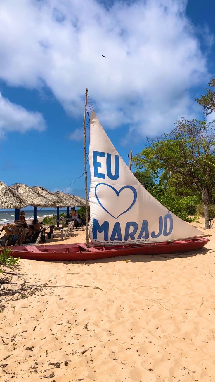 A traditional boat rests on the sandy beach, adorned with a large sail featuring the phrase 'EU ♥ MARAJO'. The shoreline is lined with gentle waves lapping at the sand, while greenery and a rustic structure are visible in the background under a bright blue sky.