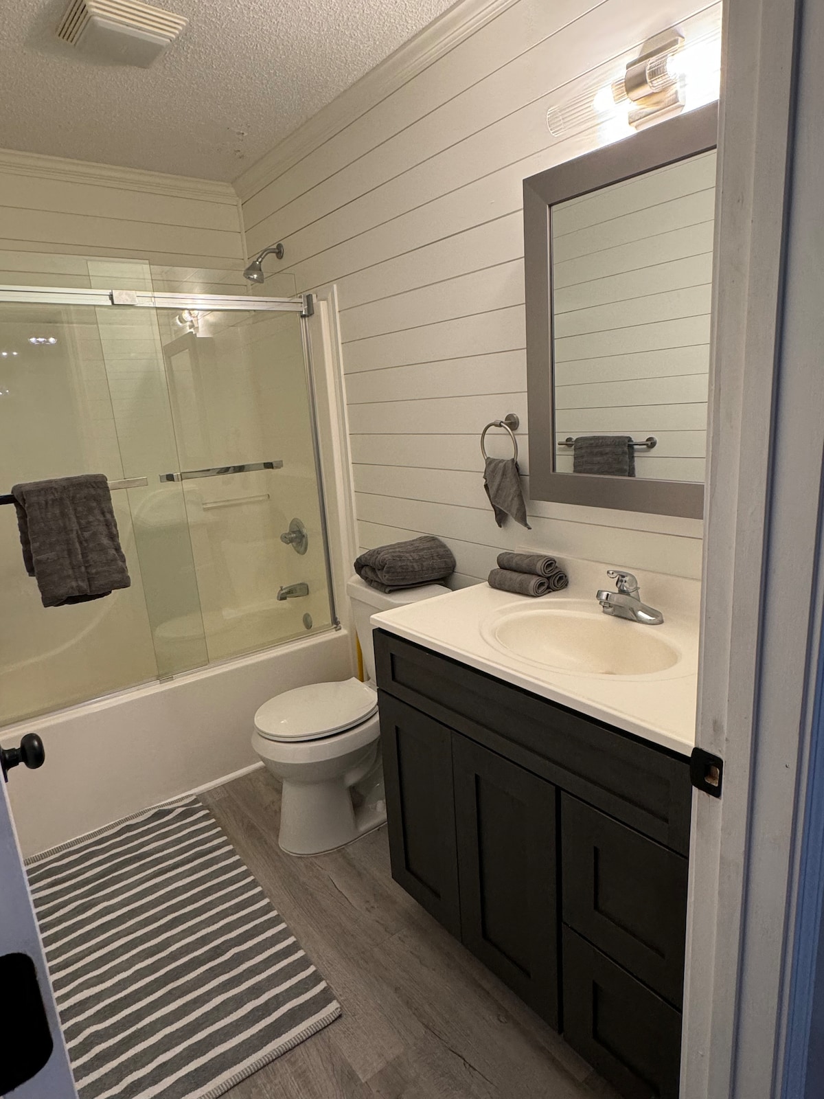 The bathroom features a modern design with a glass shower and a neutral color palette. A large mirror reflects overhead lighting, while a dark vanity contrasts with a light countertop. Towels are neatly arranged, and a gray striped rug adds texture to the floor.