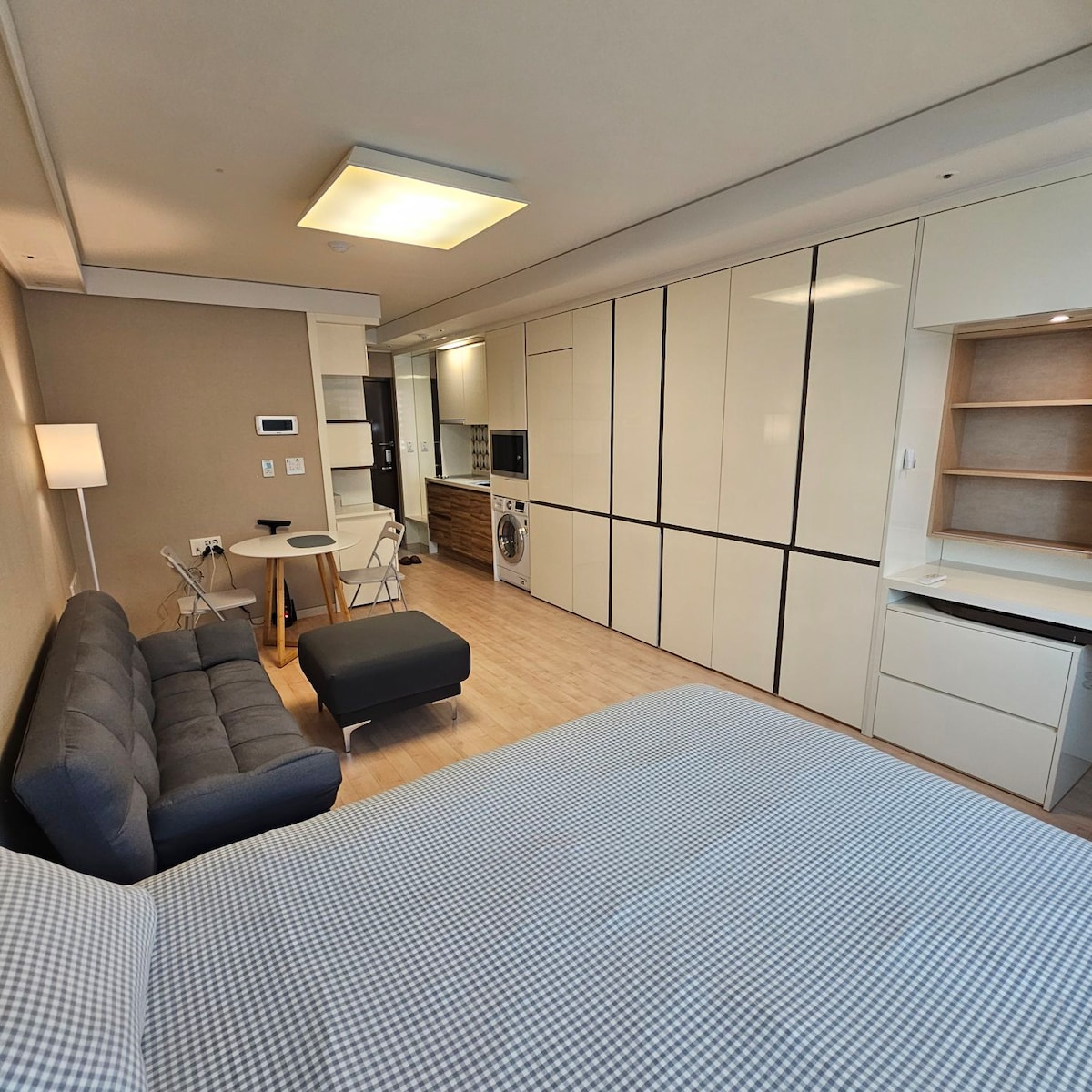 An open floor plan features a cozy seating area with a grey sofa and a small dining table for two. Light-colored cabinets and wooden accents provide ample storage. A compact kitchenette and laundry area are also visible, contributing to the functional layout of the space.