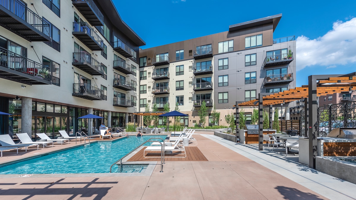 An outdoor pool area is presented, featuring lounge chairs arranged around the water. Several umbrellas provide shade, while the modern building with large windows forms a backdrop. Lush landscaping and seating areas enhance the inviting atmosphere of the poolside space.