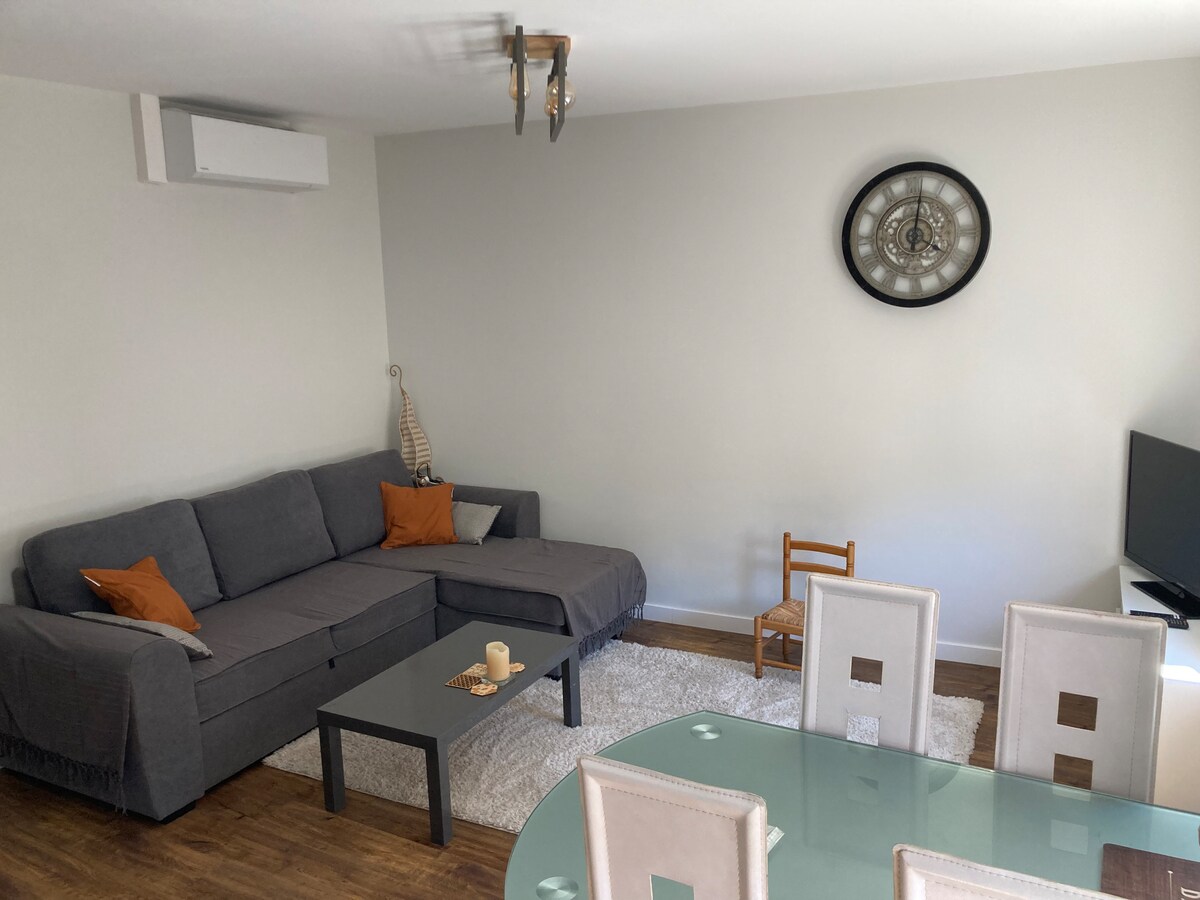 A living area features a gray sectional sofa with orange cushions and a glass coffee table. A round wall clock is mounted above, and a small wooden chair is positioned nearby. The dining area includes a glass table surrounded by light-colored upholstered chairs. An air conditioning unit is visible in the corner.
