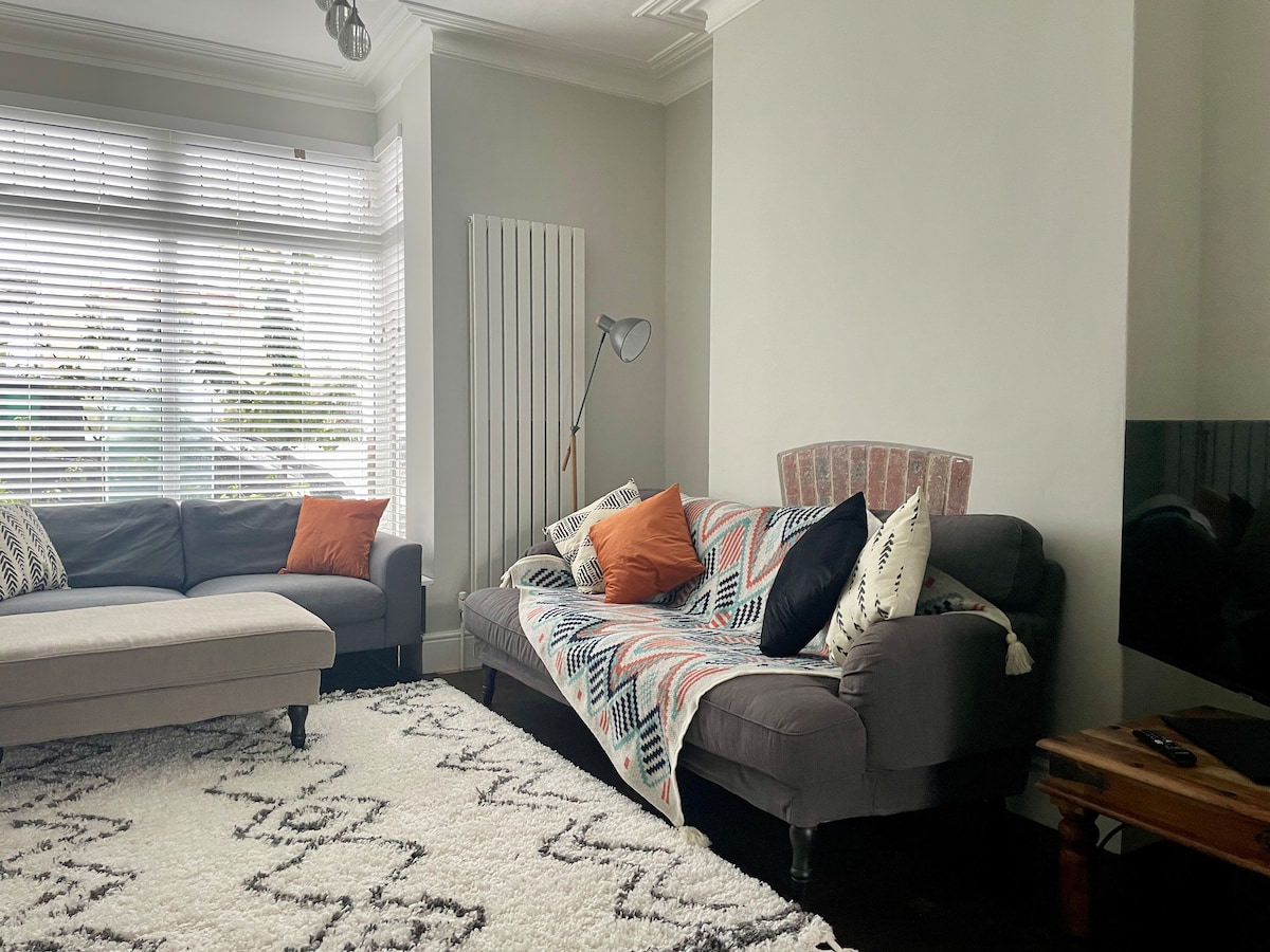The living area features a light-colored sectional sofa and a dark gray couch draped with a patterned throw blanket. An accent pillow adds warmth, while large windows provide natural light, partially covered by blinds. A plush area rug offers comfort underfoot.