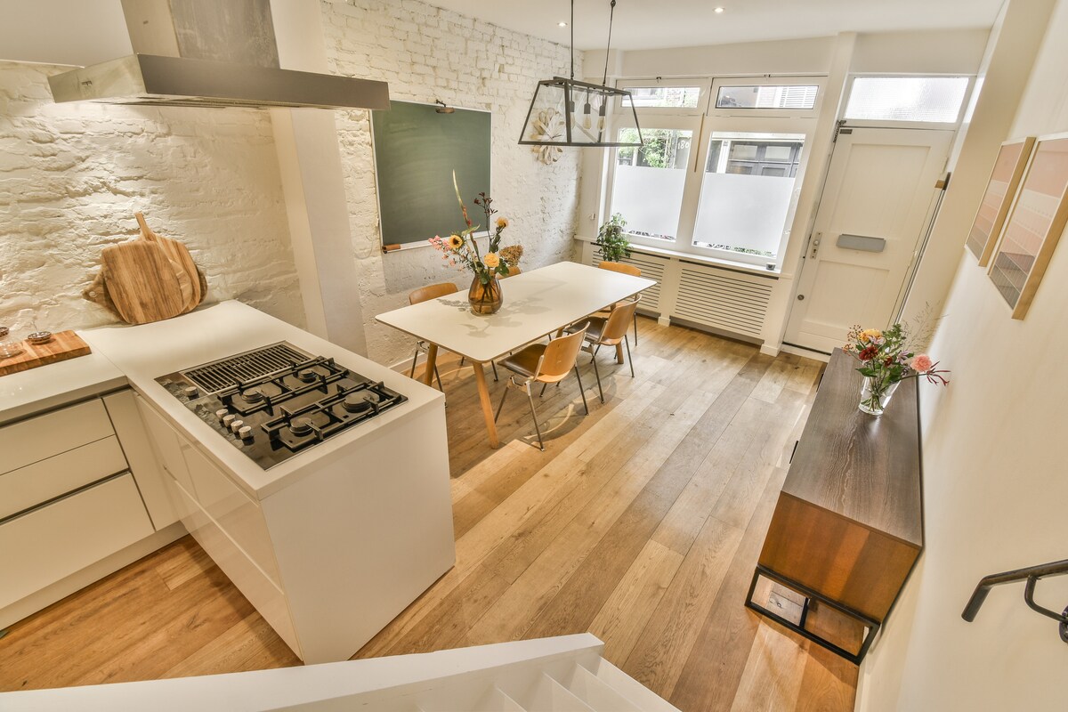 A modern kitchen is paired with a dining area, featuring a large table surrounded by wooden chairs. Light wood flooring complements white brick walls and sleek cabinetry. A large window invites natural light, while a minimalist chandelier adds subtle elegance overhead.