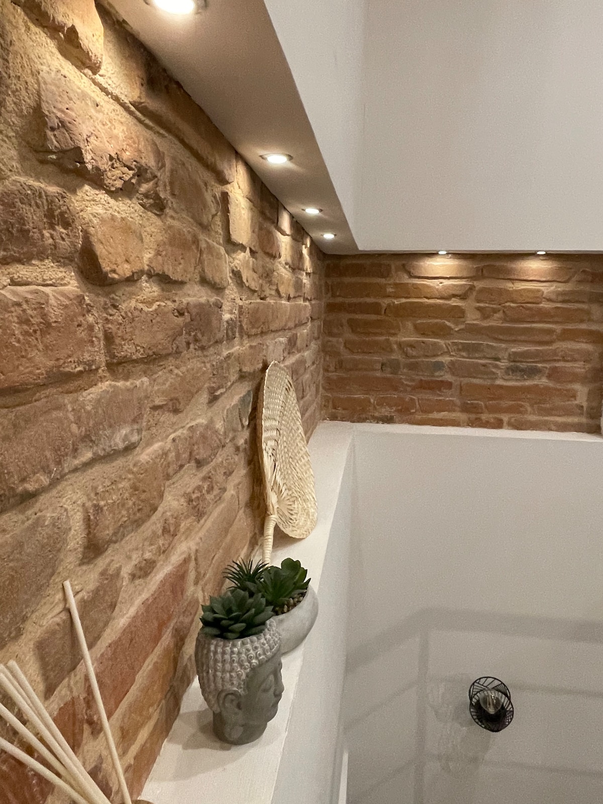 The bathroom features a white, modern tub against a backdrop of warm, exposed brick walls, which are subtly illuminated by recessed lighting. A decorative plant and a minimalist vase add a touch of greenery, creating a peaceful ambiance.