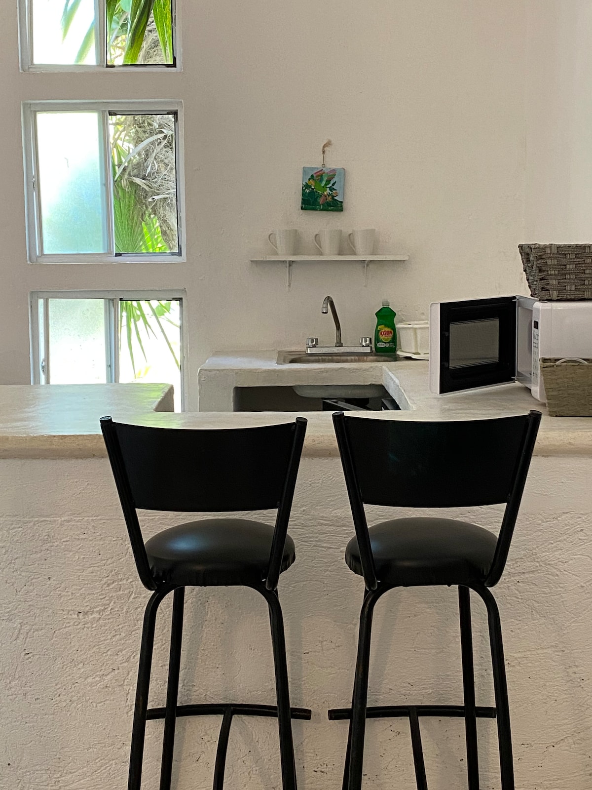 A minimalist kitchen area features a light-colored countertop with two black bar stools positioned beside it. A small sink is visible along with a microwave and a few kitchen essentials displayed on a shelf. Natural light filters in through the nearby windows, adding brightness to the space.