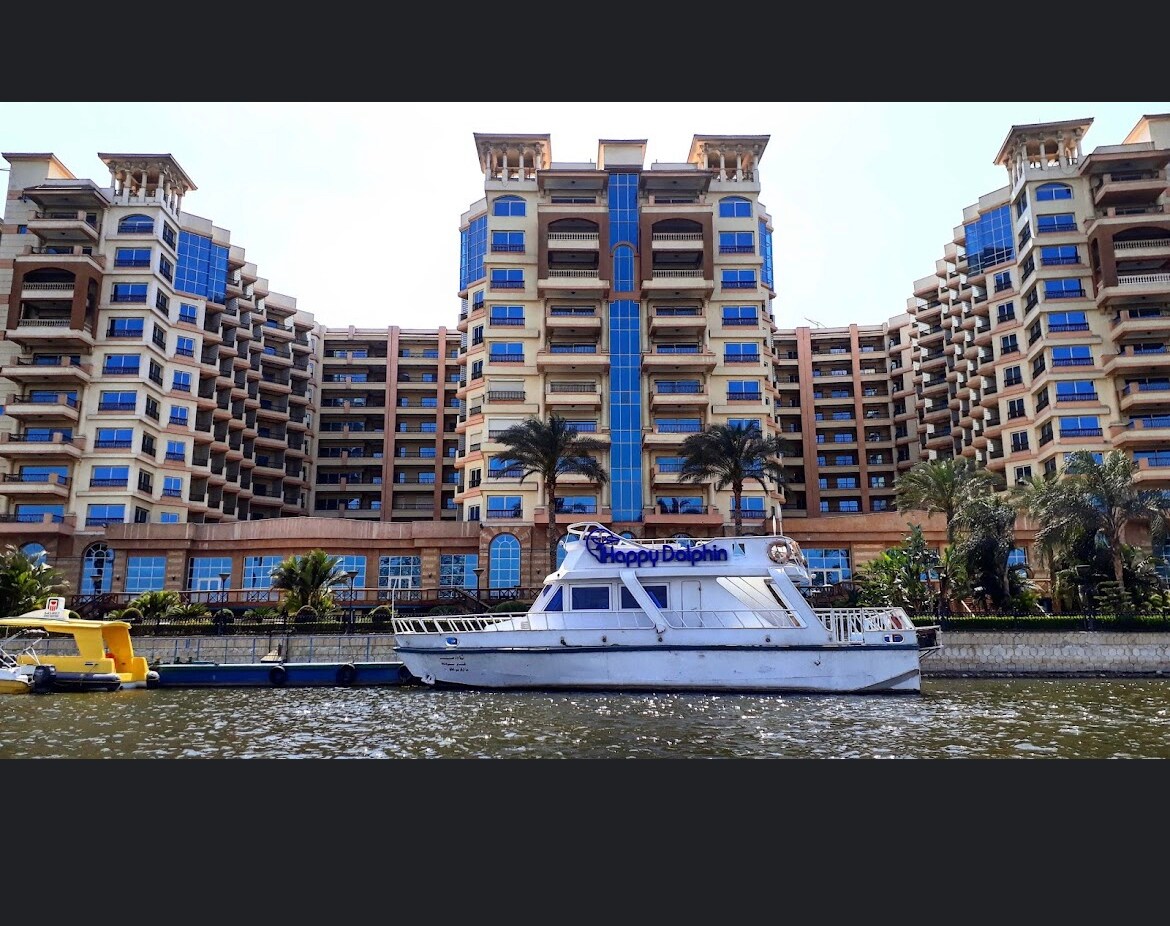 A modern waterfront building presents a spacious architectural design, featuring multiple stories with balconies and large windows. Palm trees are positioned along the water's edge, with a white boat named 'Happy Dolphin' docked nearby, enhancing the serene riverside view.