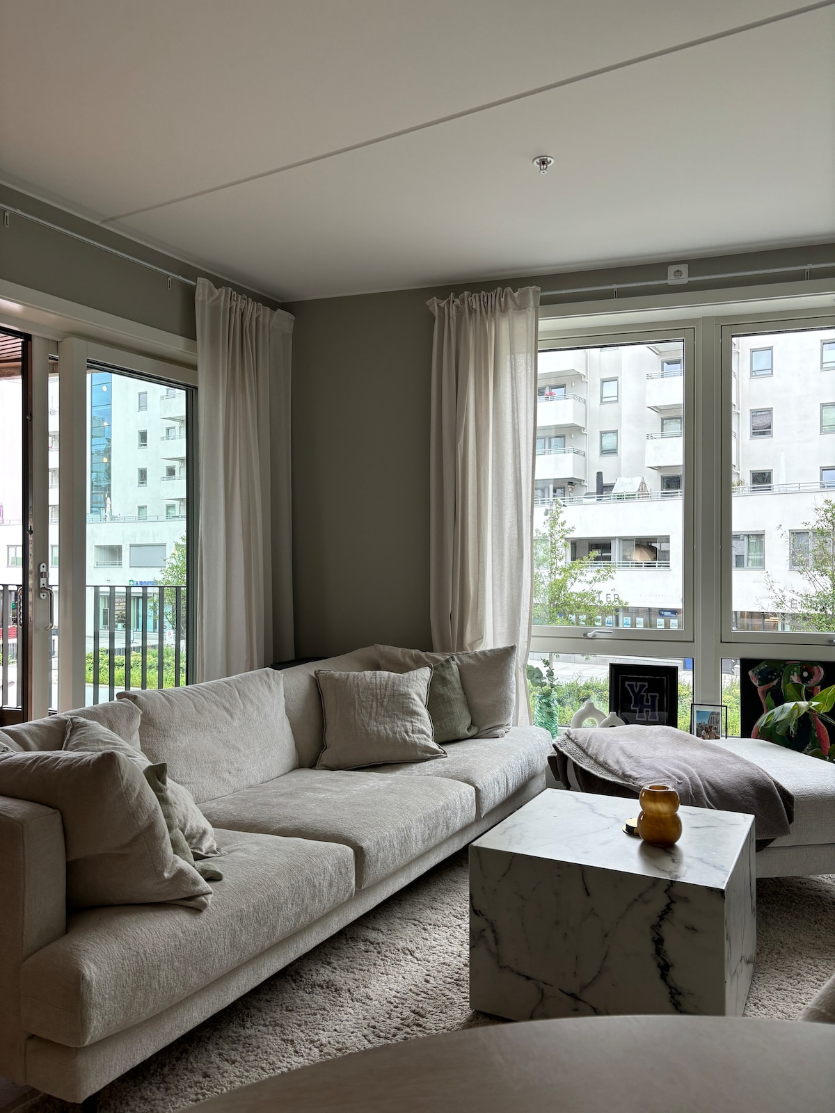 A light-filled living area features a comfortable sofa with several cushions. A marble coffee table is positioned in front, complemented by a small decorative bowl. Large windows offer expansive views of the surrounding buildings and greenery, while soft curtains frame the view.