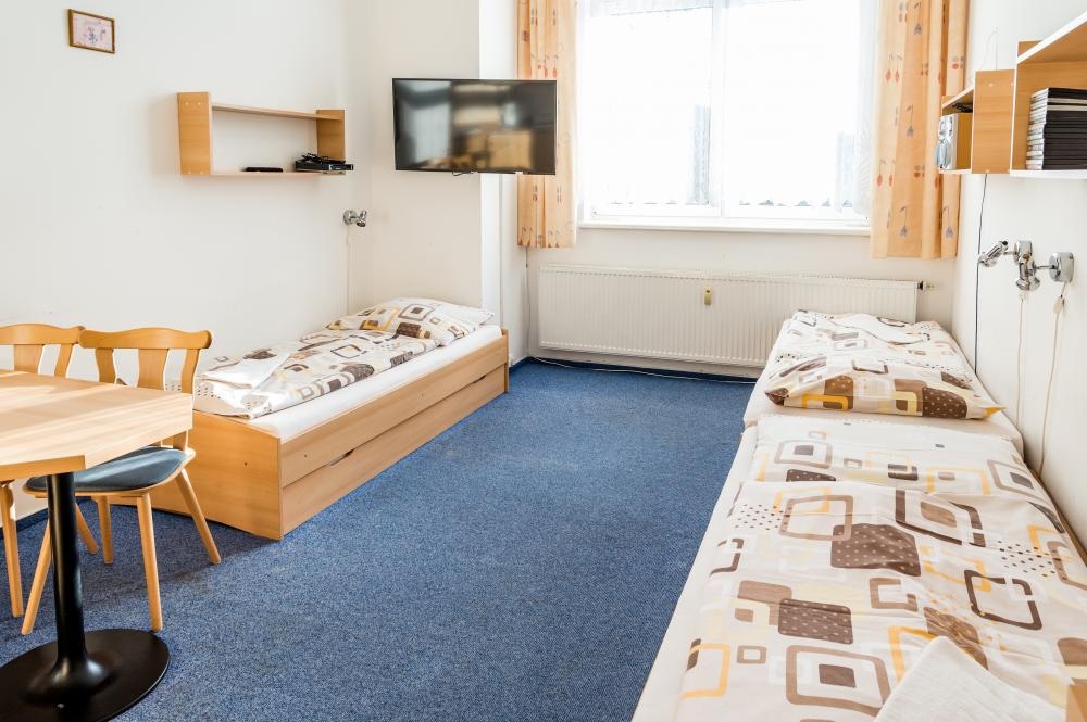 A bright room features two single beds dressed in patterned linens, positioned parallel to each other. A wooden table with two chairs is placed in the corner. A television mounted on the wall provides entertainment, while a window allows natural light to fill the space.