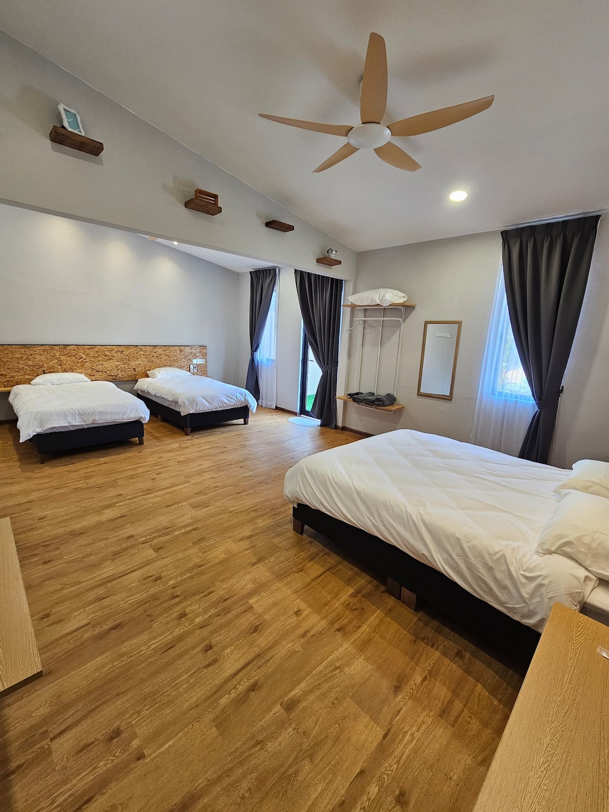 A spacious room features three beds with white bedding, arranged on dark frames. Natural light enters through a large window dressed with dark curtains. A ceiling fan adds to the airiness, while wooden accents adorn the walls, enhancing the room's modern aesthetic.