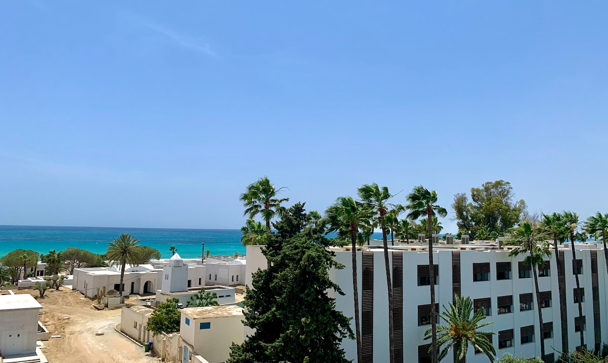 A view of the coastline features gentle waves and palm trees lining the shore. The surrounding buildings, characterized by white facades, provide a contrast against the clear blue sky. The natural landscape evokes a sense of tranquility and proximity to the beach.