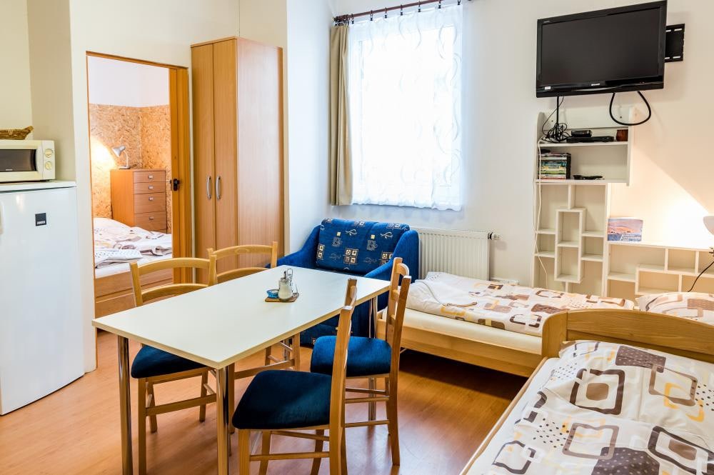 An open living area is depicted, featuring a small dining table with four blue chairs, a sofa, and a single bed. A television is mounted on the wall, and a window allows natural light to enter, complemented by light-colored curtains. Storage units are visible in the background.