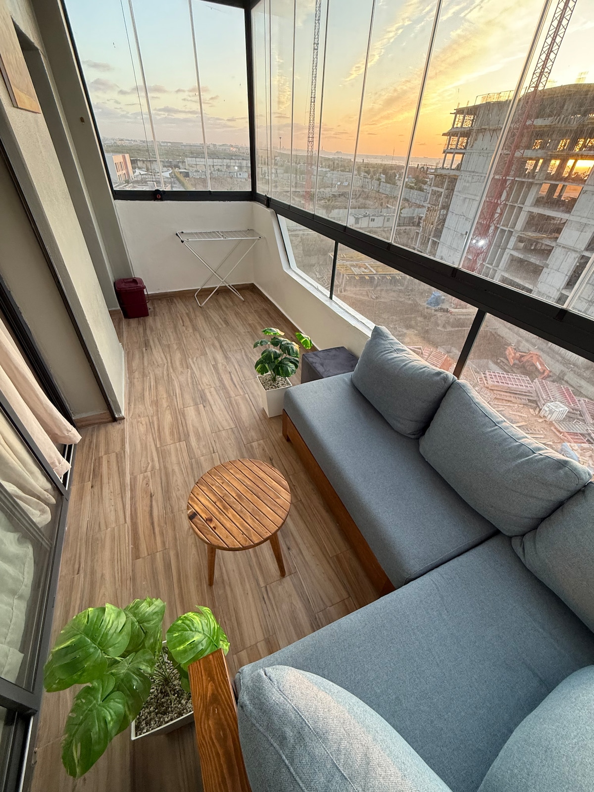 A cozy balcony is furnished with a comfortable, spacious sofa, a small wooden table, and a potted plant. Large windows offer a view of the sunset, while warm light fills the space. The flooring features wood-like tiles, enhancing the inviting ambiance.