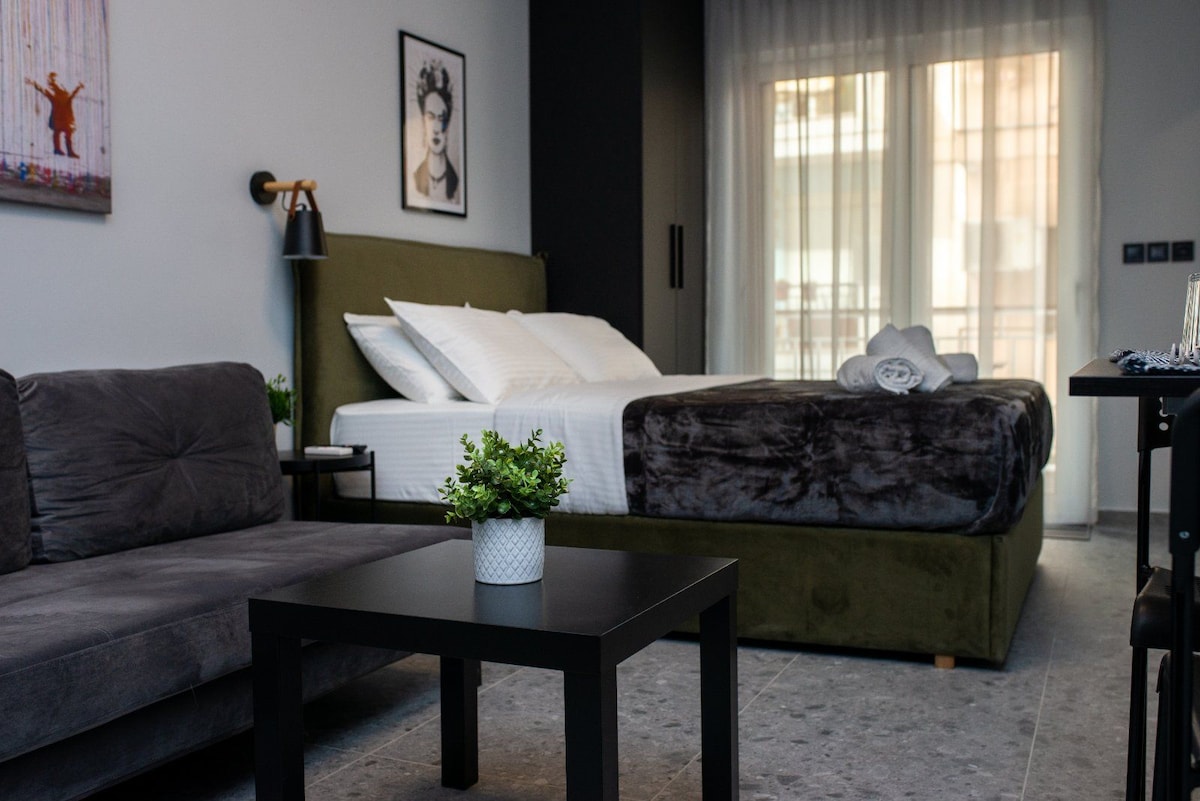 A studio space showcases a spacious bed dressed in soft linens, accompanied by a cozy gray sofa and a modern black coffee table. Natural light filters through sheer curtains, illuminating a decorative wall art piece and a small potted plant on the table.