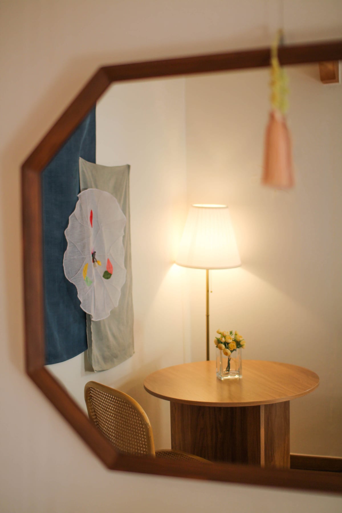 A small round table is positioned beneath a soft-lit lamp, creating a cozy corner. A decorative wall hanging featuring floral designs is mounted behind the table. A vase with fresh flowers adds a touch of nature to the setting, reflected in the octagon-shaped mirror.