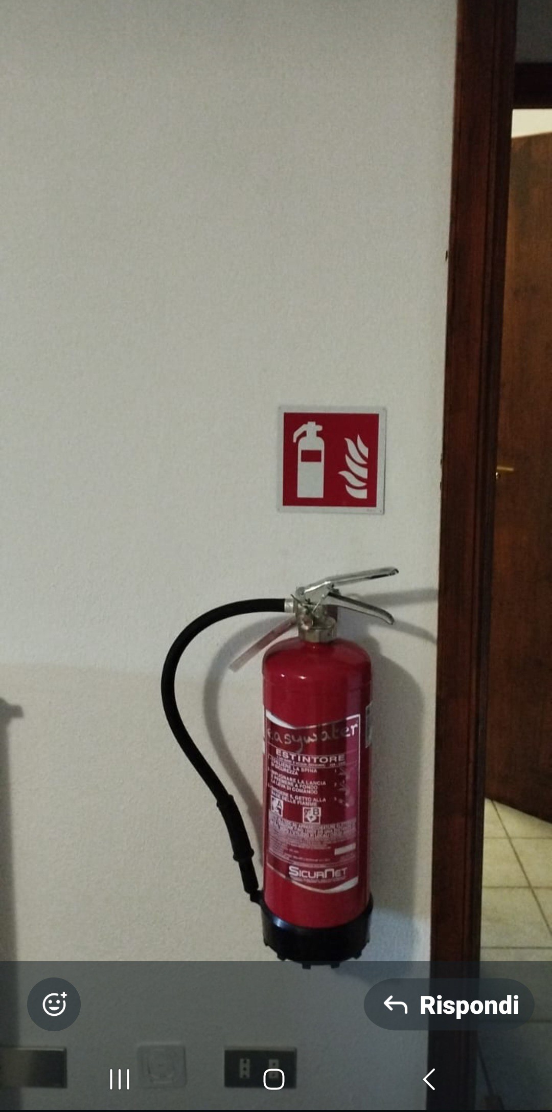A red fire extinguisher is mounted on a wall next to a fire safety sign. The sign features a fire symbol, clearly indicating fire safety measures. The background shows a neutral-colored wall, contributing to the safety-focused environment.