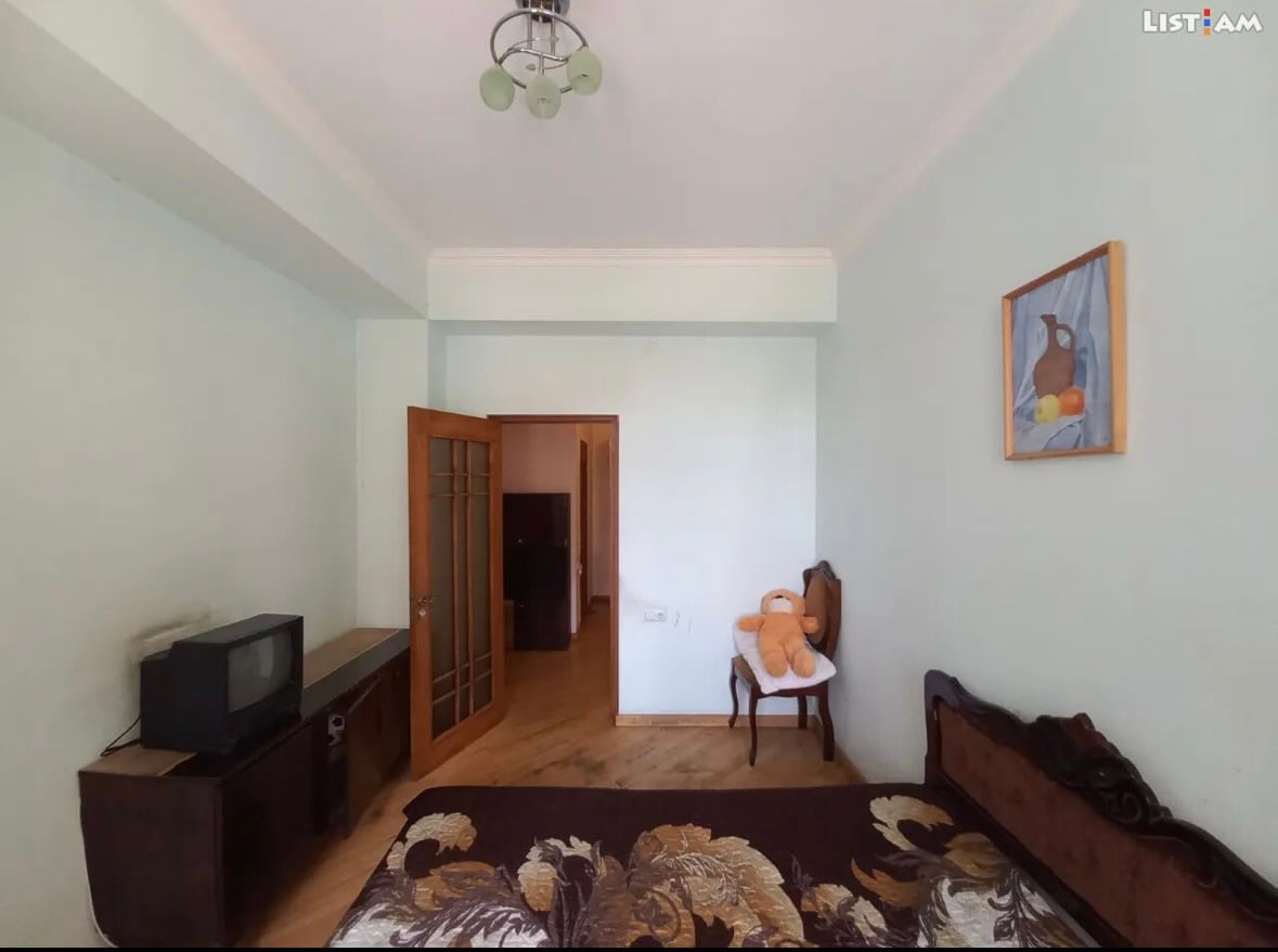 A light-coloured bedroom features a spacious bed adorned with a floral-patterned blanket. A vintage wooden chair is positioned beside the bed. A television stands on a wooden console against the wall, and a painting of fruit decorates the wall beside the door.