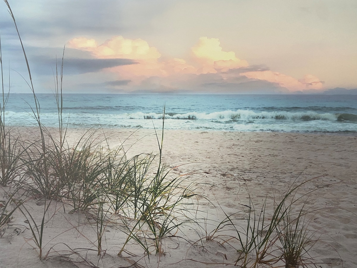 The image features a serene beach scene with gentle waves lapping against soft sand. Grasses are visible in the foreground, swaying slightly in the breeze. Soft clouds and pastel colors decorate the sky, providing a calming backdrop to the tranquil ocean landscape.