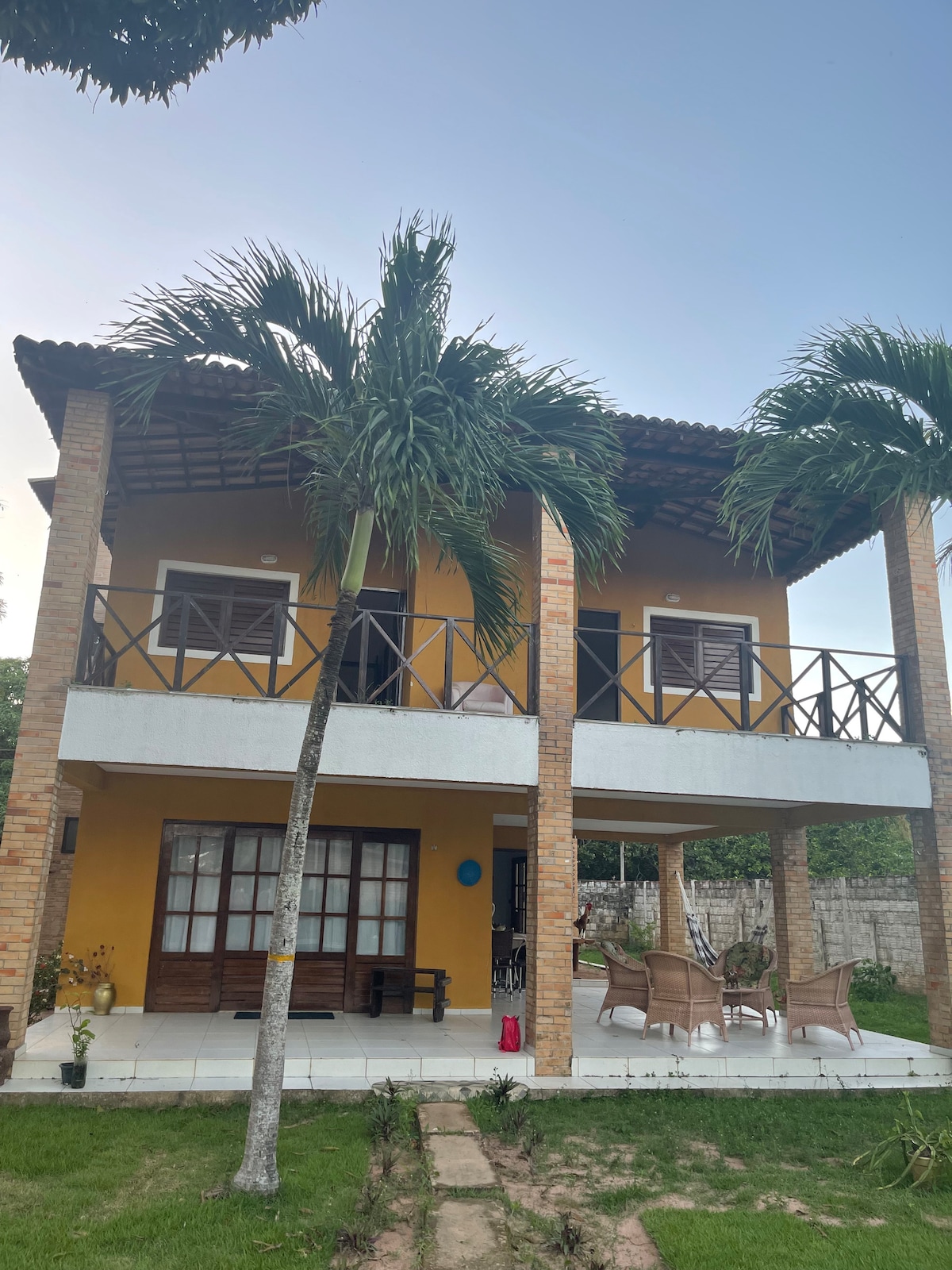 A two-story house is set amidst palm trees, featuring a yellow exterior and a covered balcony. The ground level showcases a spacious patio with outdoor seating, while large windows provide ample natural light. A well-maintained lawn leads to a stone pathway.