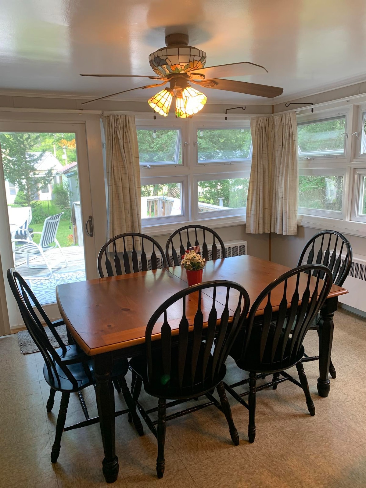 A dining area features a wooden table surrounded by six black chairs. Natural light enters through multiple windows with light curtains, while a ceiling fan hangs above. A small potted plant is placed at the center of the table, adding a touch of greenery.