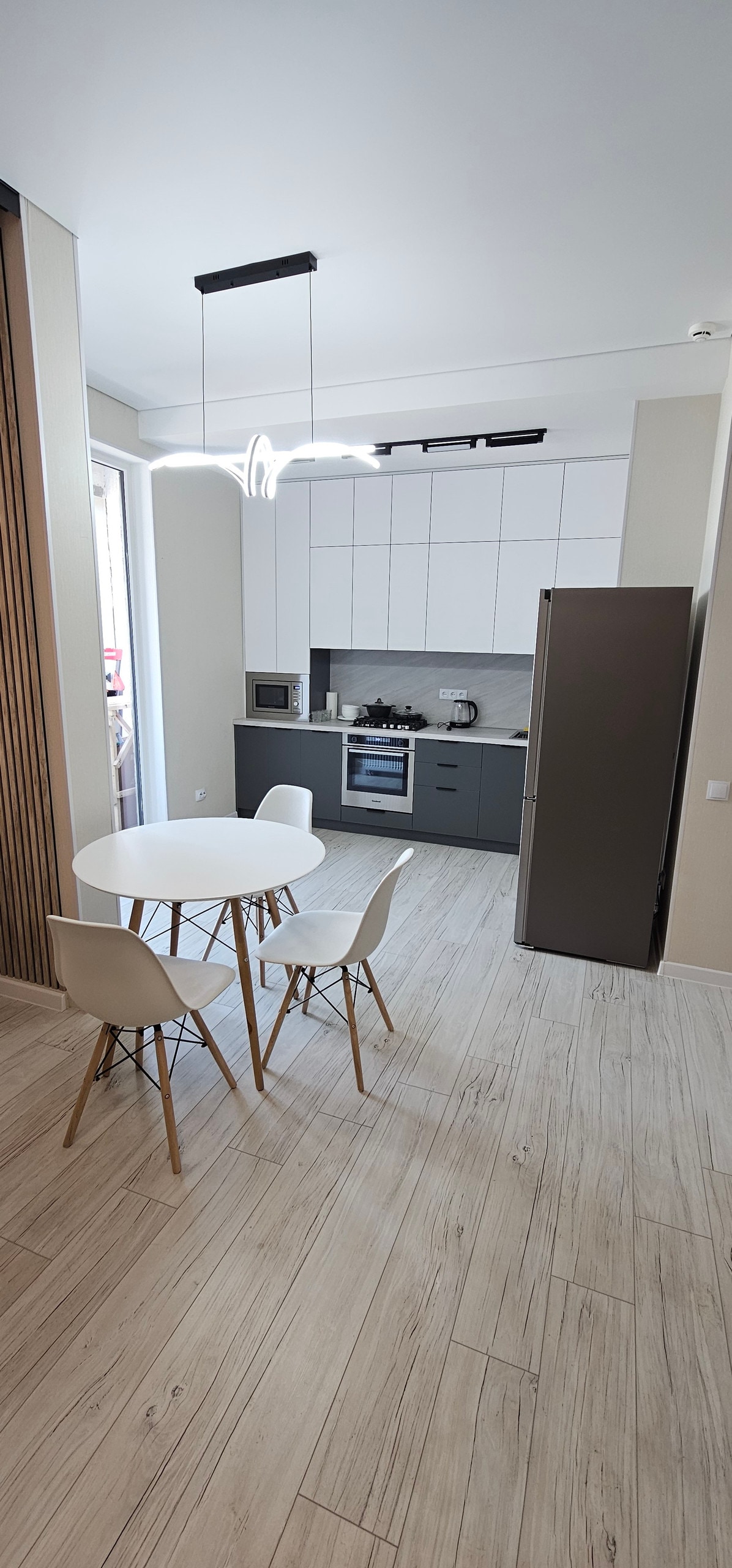 A modern kitchen area is equipped with sleek cabinetry and appliances, featuring a stove and microwave. A round dining table with four white chairs is centrally positioned. Natural light enters through a large window, illuminating the light wood flooring.