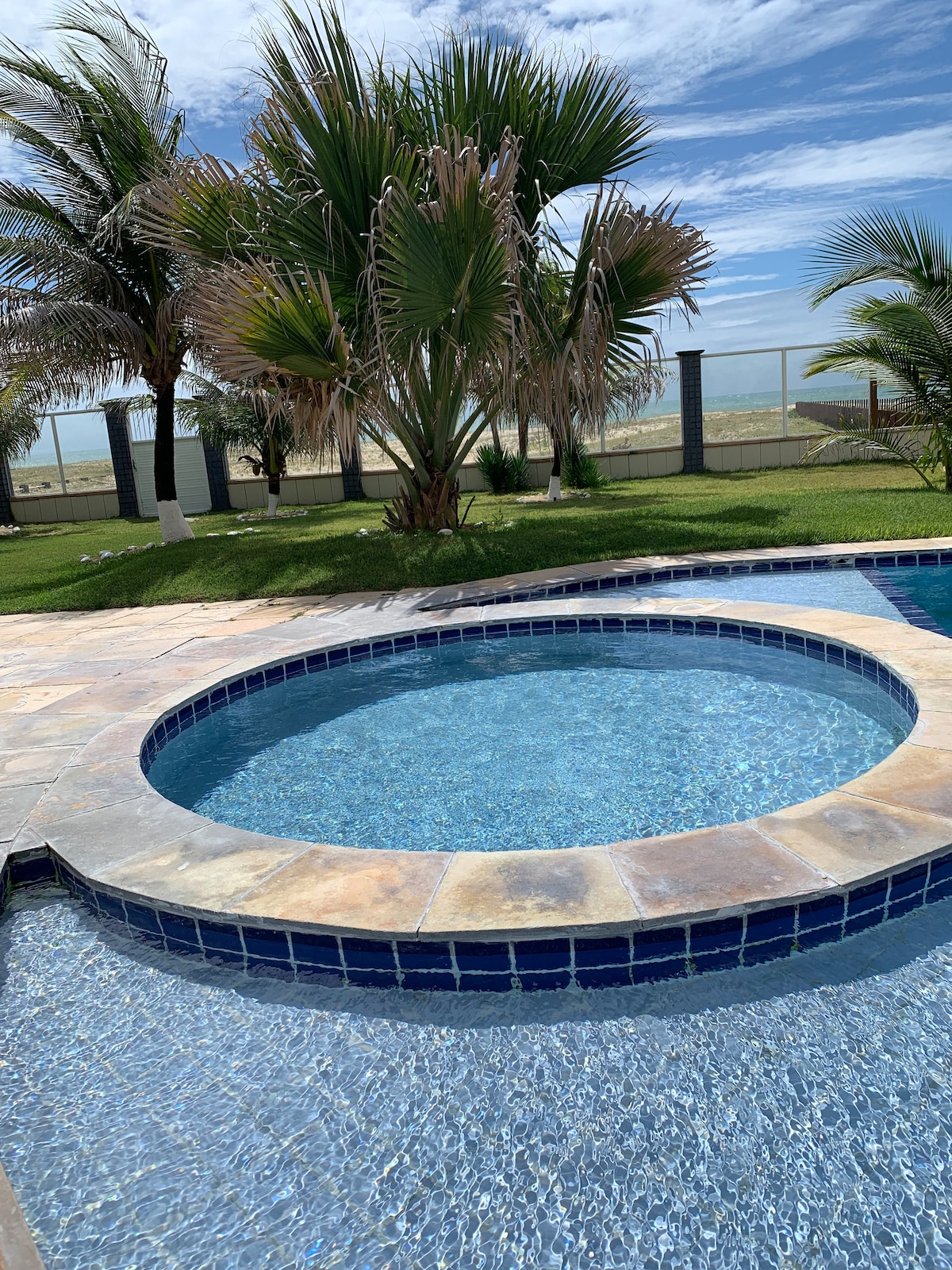 A well-maintained swimming pool area features a circular hot tub alongside a sparkling blue pool. Lush palm trees surround the space, contributing to a relaxing outdoor environment. A clear view of the beach is visible in the background, enhancing the serene atmosphere.