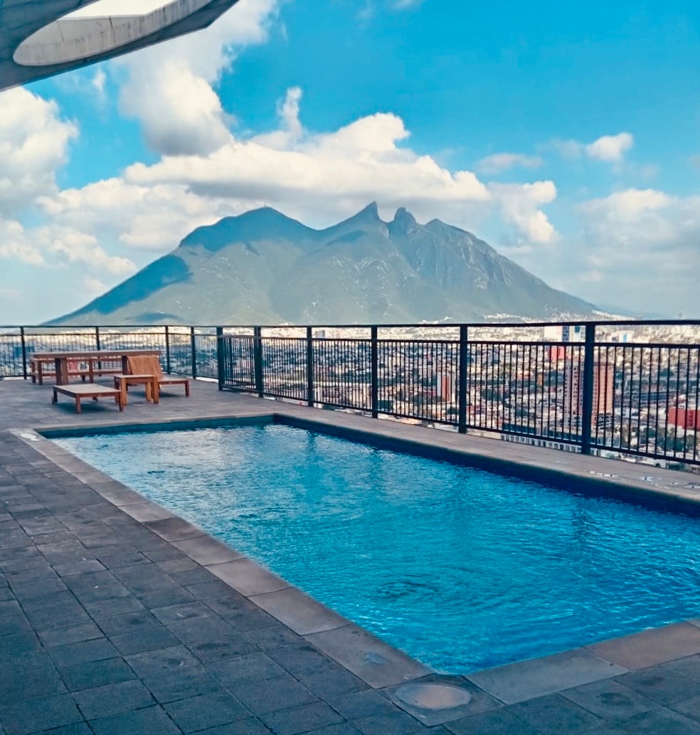An outdoor pool is positioned on a rooftop terrace, offering views of the surrounding cityscape and the prominent Cerro de la Silla mountain. Lounge chairs are placed beside the pool, and the sky is dotted with clouds.